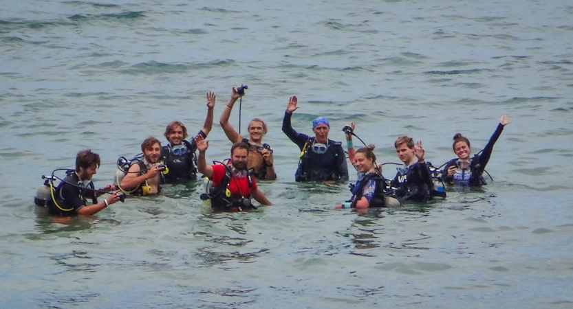 A group of people wearing SCUBA gear stand in waist-deep water and wave at the camera. 